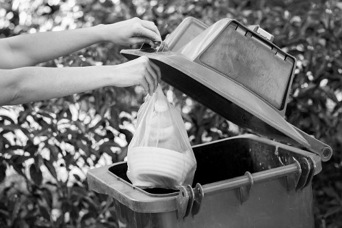 A pair of human hands are shown lifting the lid of a large outdoor rubbish bin, revealing a black plastic bag inside. The bin is situated on a paved area surrounded by dense shrubbery, with leafy branches in the background. The bin itself is made of durable plastic with a textured surface and a closed, hinged lid. The black bag appears to contain household waste, and the scene is set in natural daylight, indicating daytime. This image depicts typical waste collection or rubbish disposal activity consistent with services provided by Garden Clearance Finsbury Park, focusing on waste removal and rubbish management in a residential or garden setting within the Finsbury Park area.