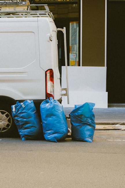 A white cargo van parked on a city street with its rear door open, revealing a small loading area. On the ground beside the van are three large, blue, plastic rubbish bags filled with waste materials,