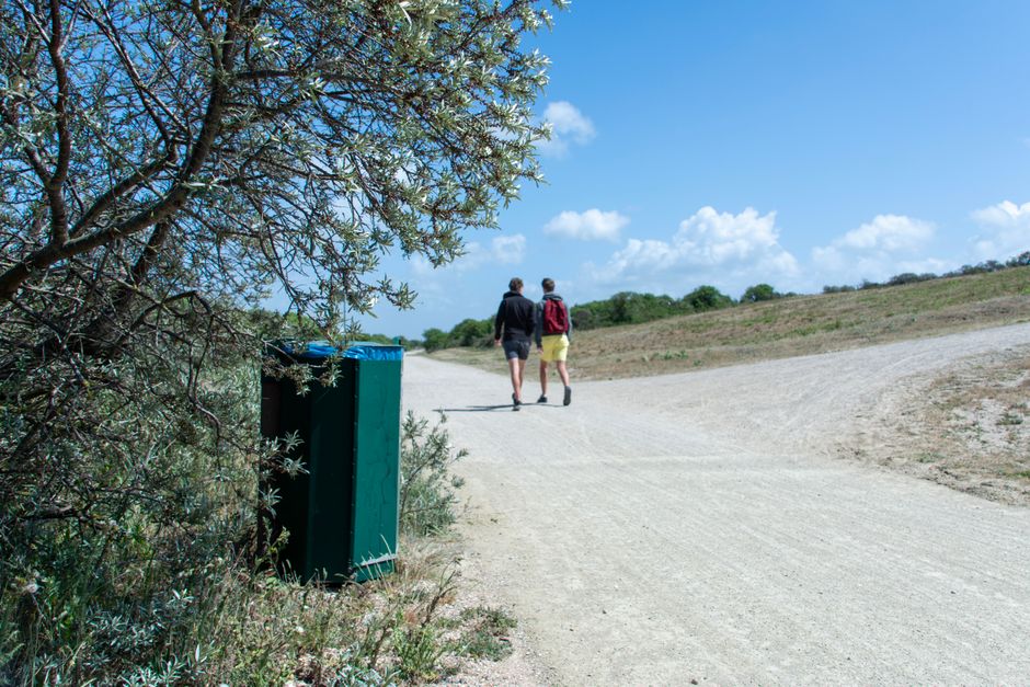 A wide, unpaved gravel path runs through a natural outdoor setting under a bright blue sky with scattered white clouds. In the foreground, a green plastic rubbish bin with a textured surface is partia